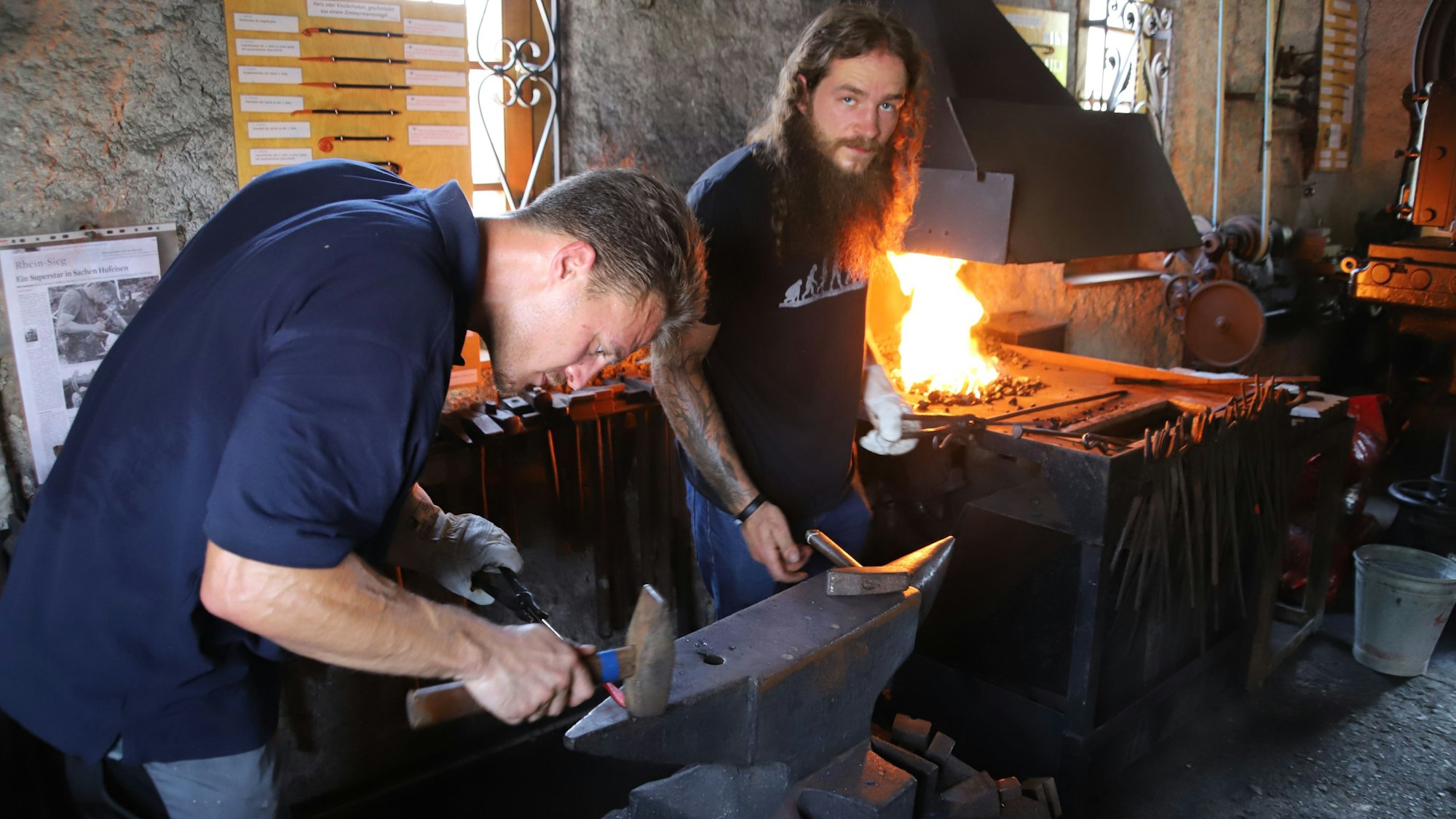 Ein Mann schlägt im Schmiedemuseum Niederkassel-Lülsdorf mit einem Hammer auf einen Amboss, im Hintergrund lodert ein Feuer in einer Esse.