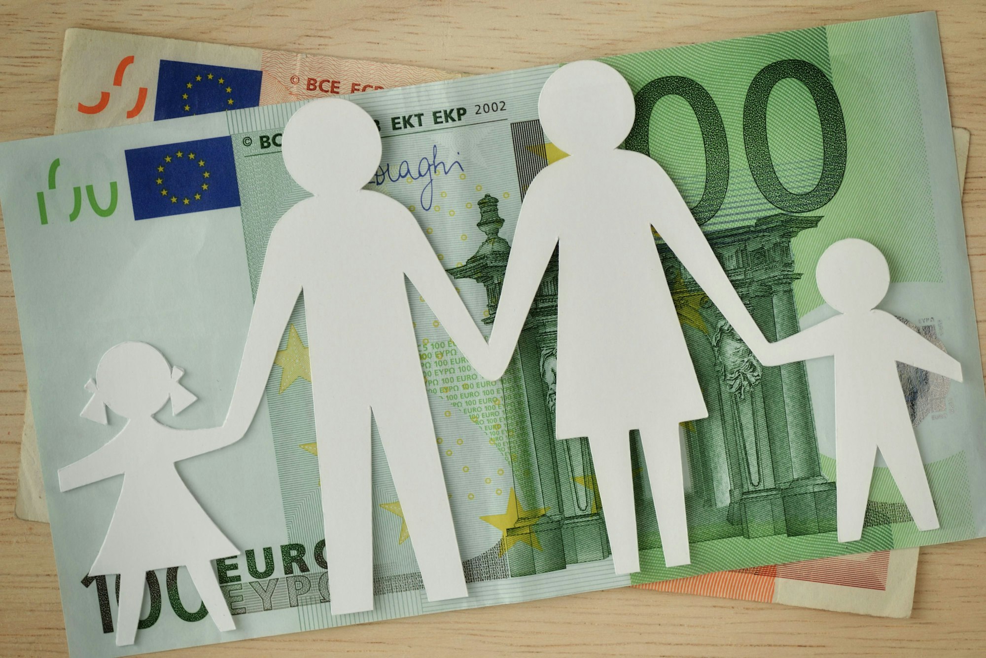 Auf einem 100-Euro-Schein, unter dem ein 50-Euro-Schein sichtbar wird, befindet sich eine aus Papier ausgeschnittene Familie, bestehend aus Vater, Mutter und zwei Kindern.