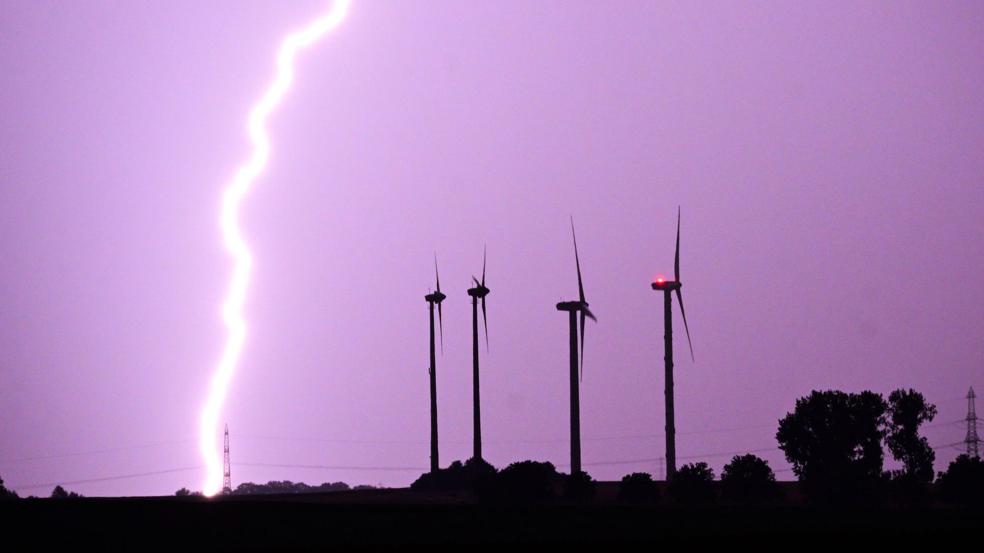 Ein Blitz entlädt sich während eines Gewitters hinter Windrädern. Der Himmel ist lila gefärbt. (Symbolbild)