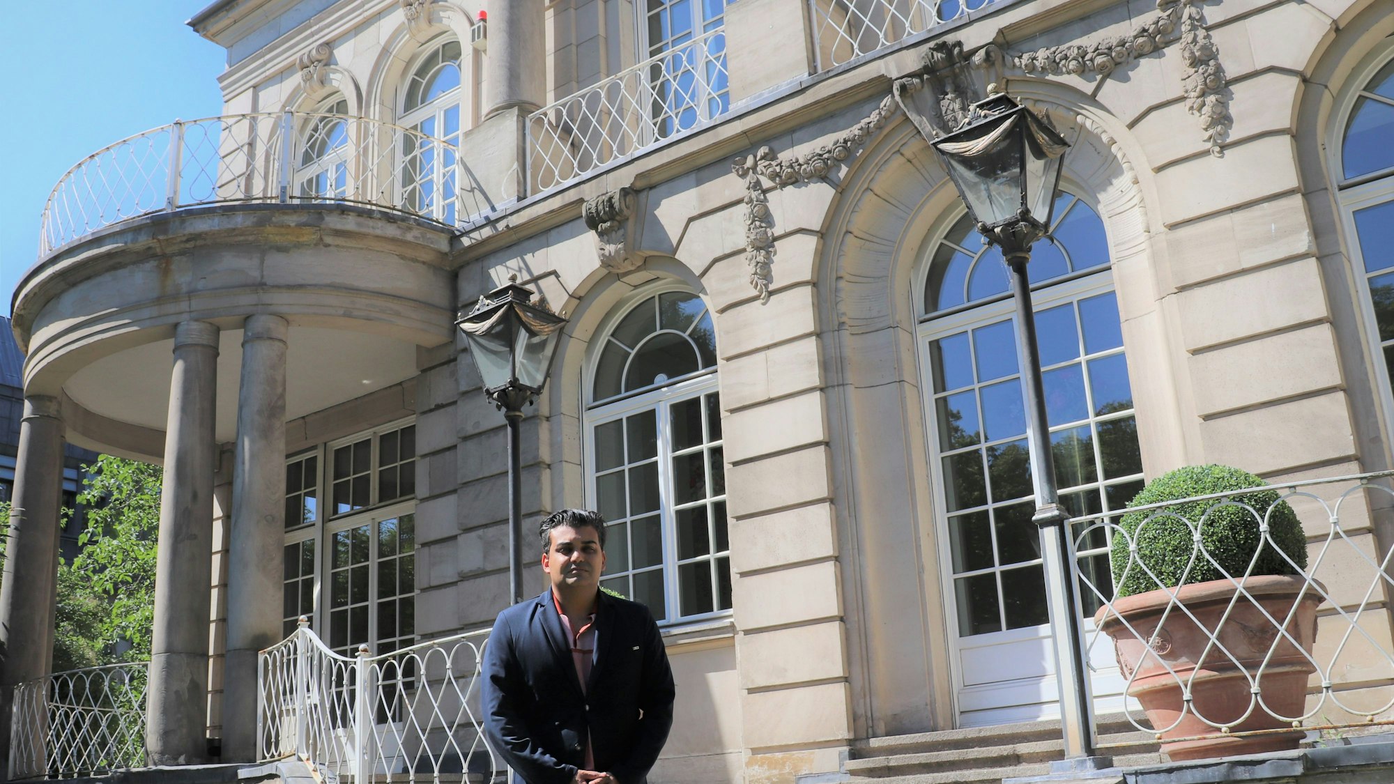 Ein Mann mit dunklem Haar und blauem Jackett steht vor einer großen Villa mit Säulen und Terrassentüren mit Rundbögen.