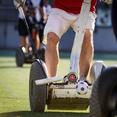 Zu sehen sind die Beine eines Mannes, der auf einem Segway steht und einen Polo-Schläger in der Hand hält.