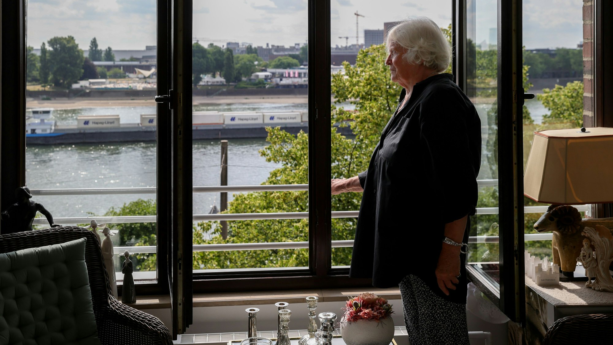 Eine weißhaarige Frau steht am Fenster und betrachtet den Rhein.