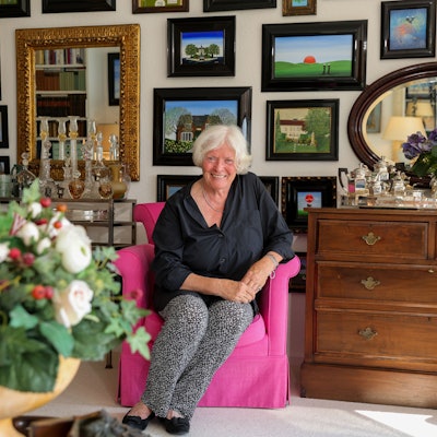Eine weißhaarige Frau sitzt in einem pinken Sessel. Dahinter hängen Bilder.