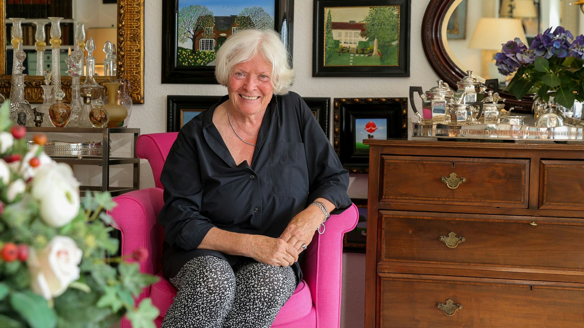 Eine weißhaarige Frau sitzt in einem pinken Sessel. Dahinter hängen Bilder.