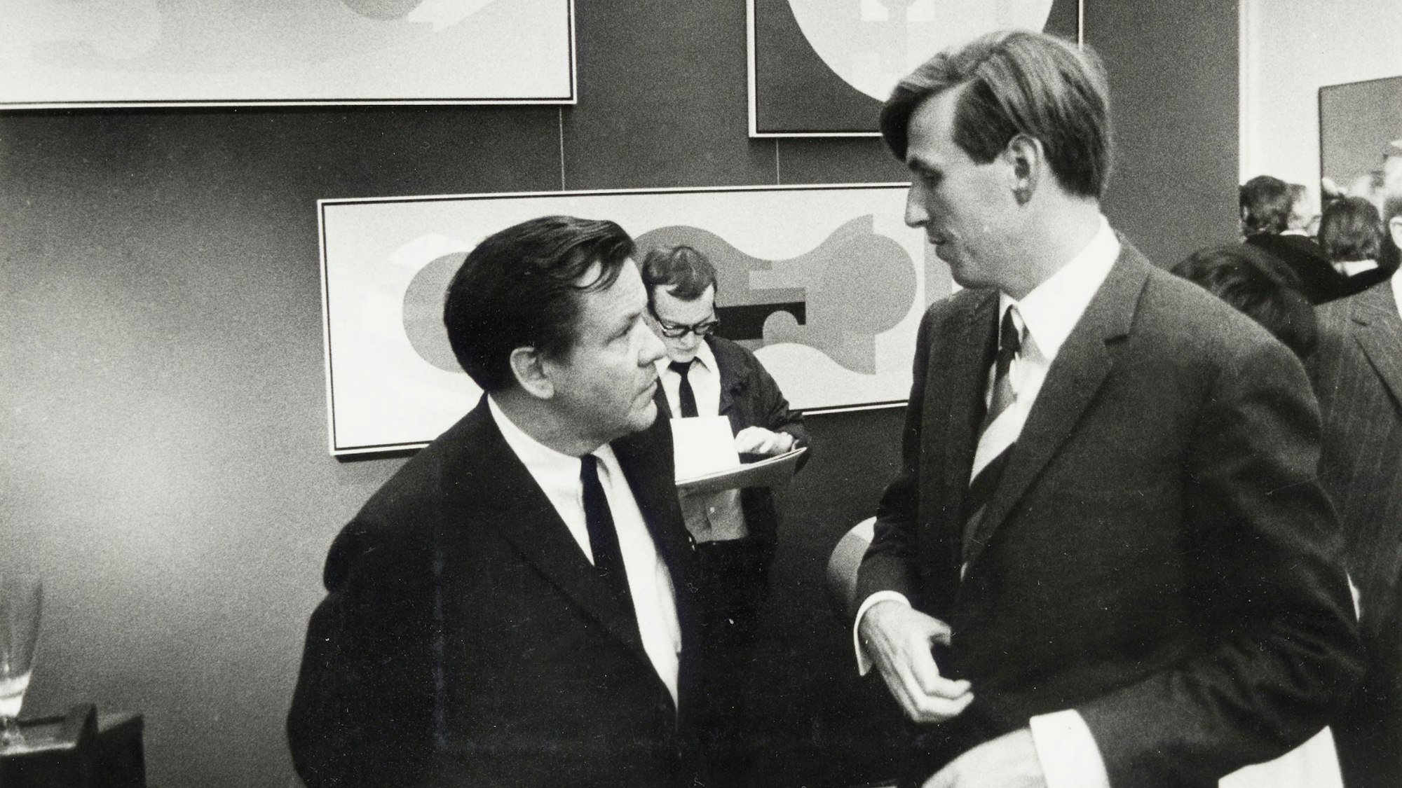 Zwei Männer in Anzügen unterhalten sich auf einer Kunstmesse.