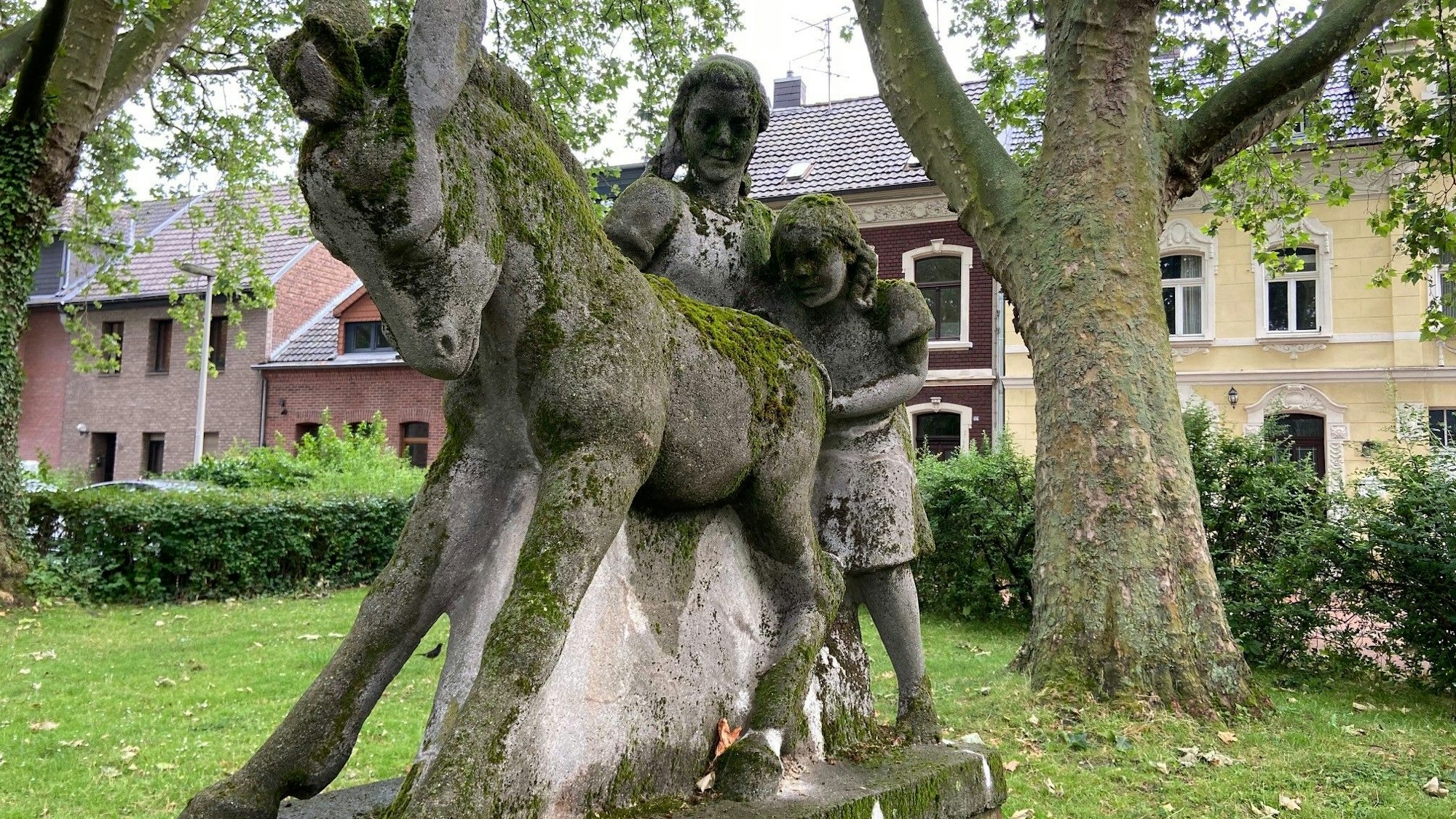 Das Foto zeigt zwei Mädchen, die sich sich mühen, einen störrischen Esel voranzutreiben. Die Steinfiguren sind moosbewachsen.