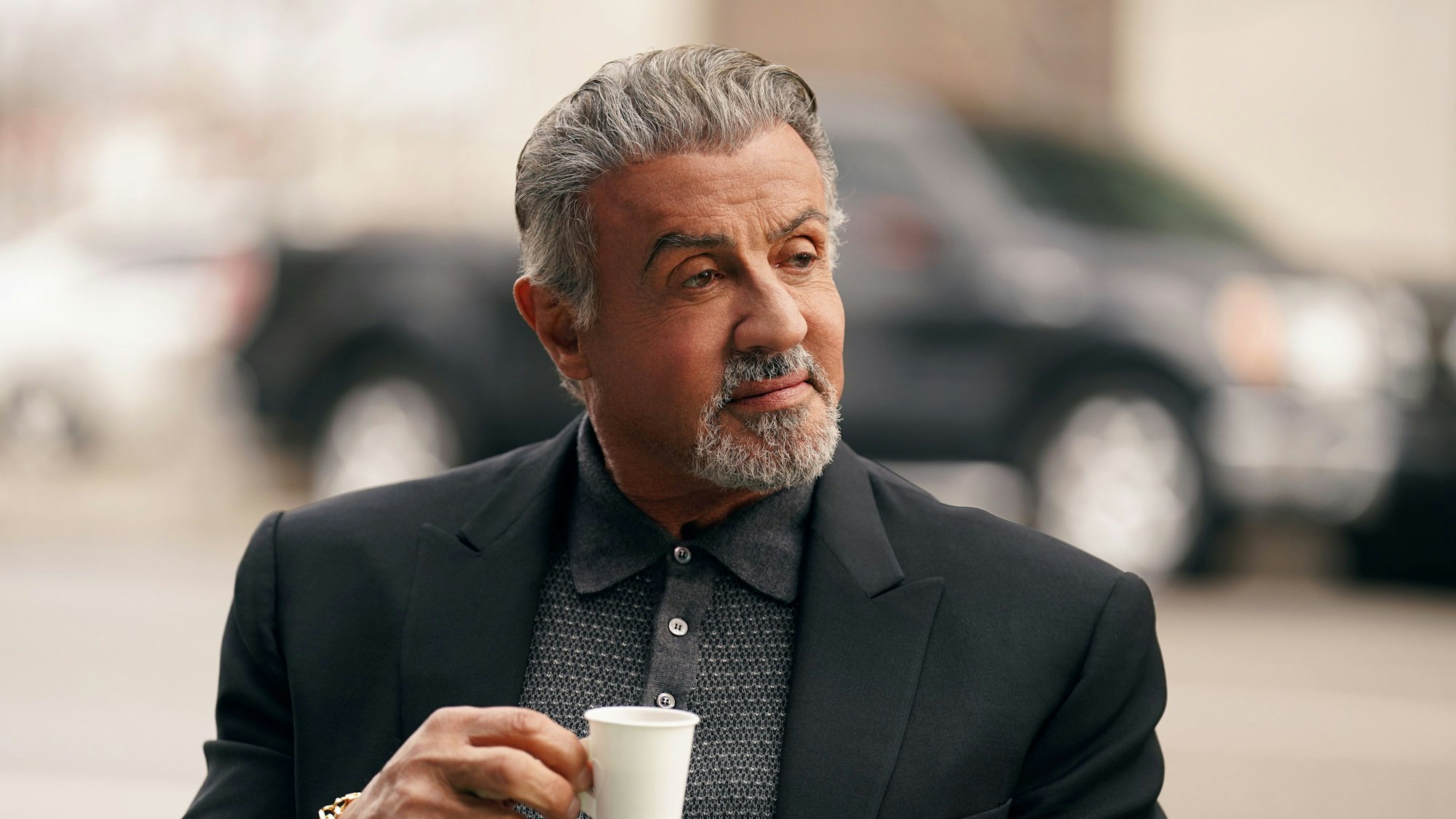 Sylvester Stallone hält einen Kaffee in der Hand und guckt zur Seite.