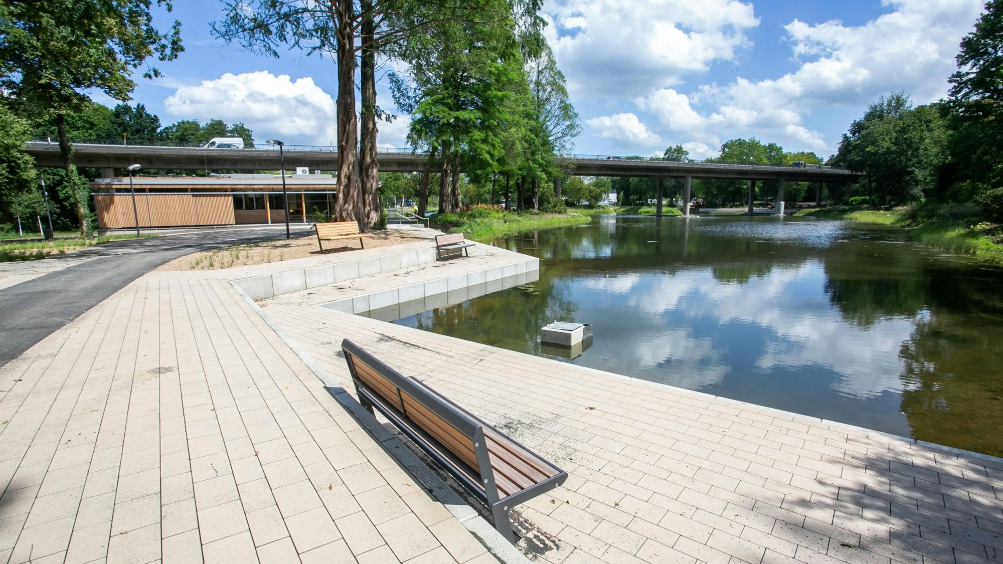 Zu sehen ist eine Sitzbank am Ufer des Teichs, im Hintergrund das neue Seecafé.