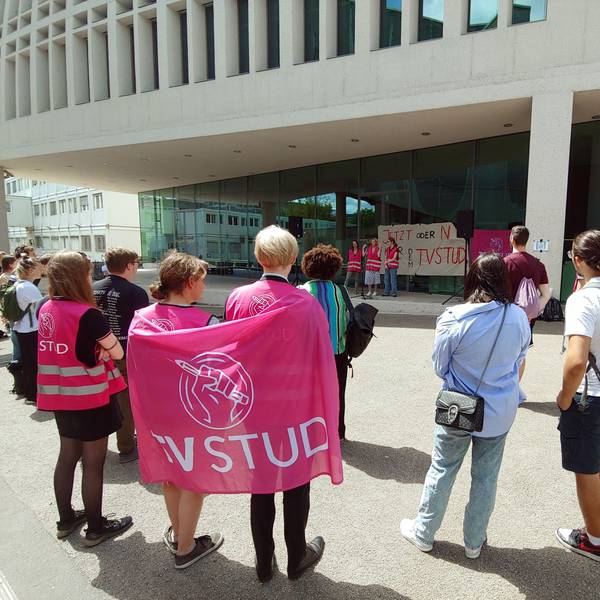 Menschen, die sich mit pinken Warnwesten und Protestschildern vor einem Gebäude mit einer Glasfassade versammelt haben