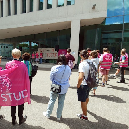 Menschen, die sich mit pinken Warnwesten und Protestschildern vor einem Gebäude mit einer Glasfassade versammelt haben