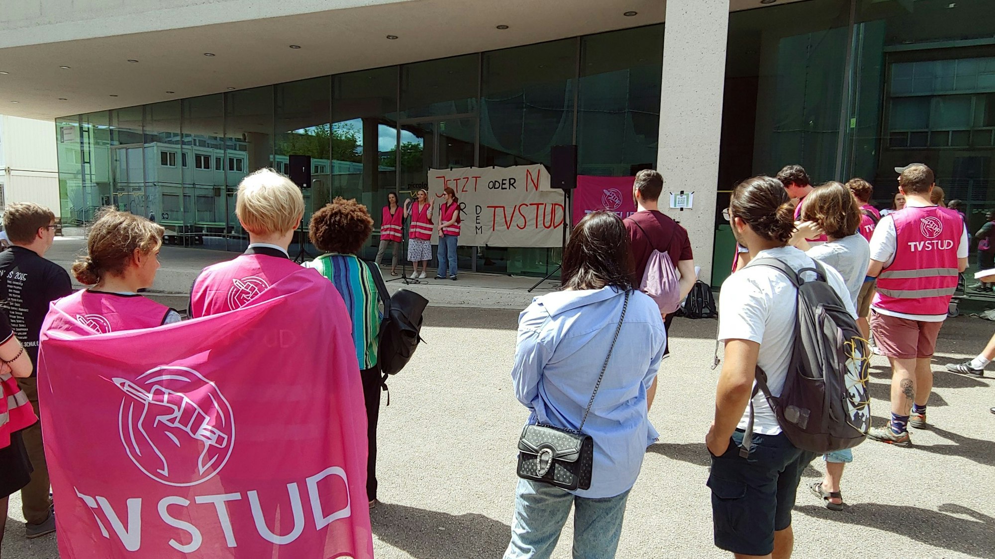 Menschen, die sich mit pinken Warnwesten und Protestschildern vor einem Gebäude mit einer Glasfassade versammelt haben