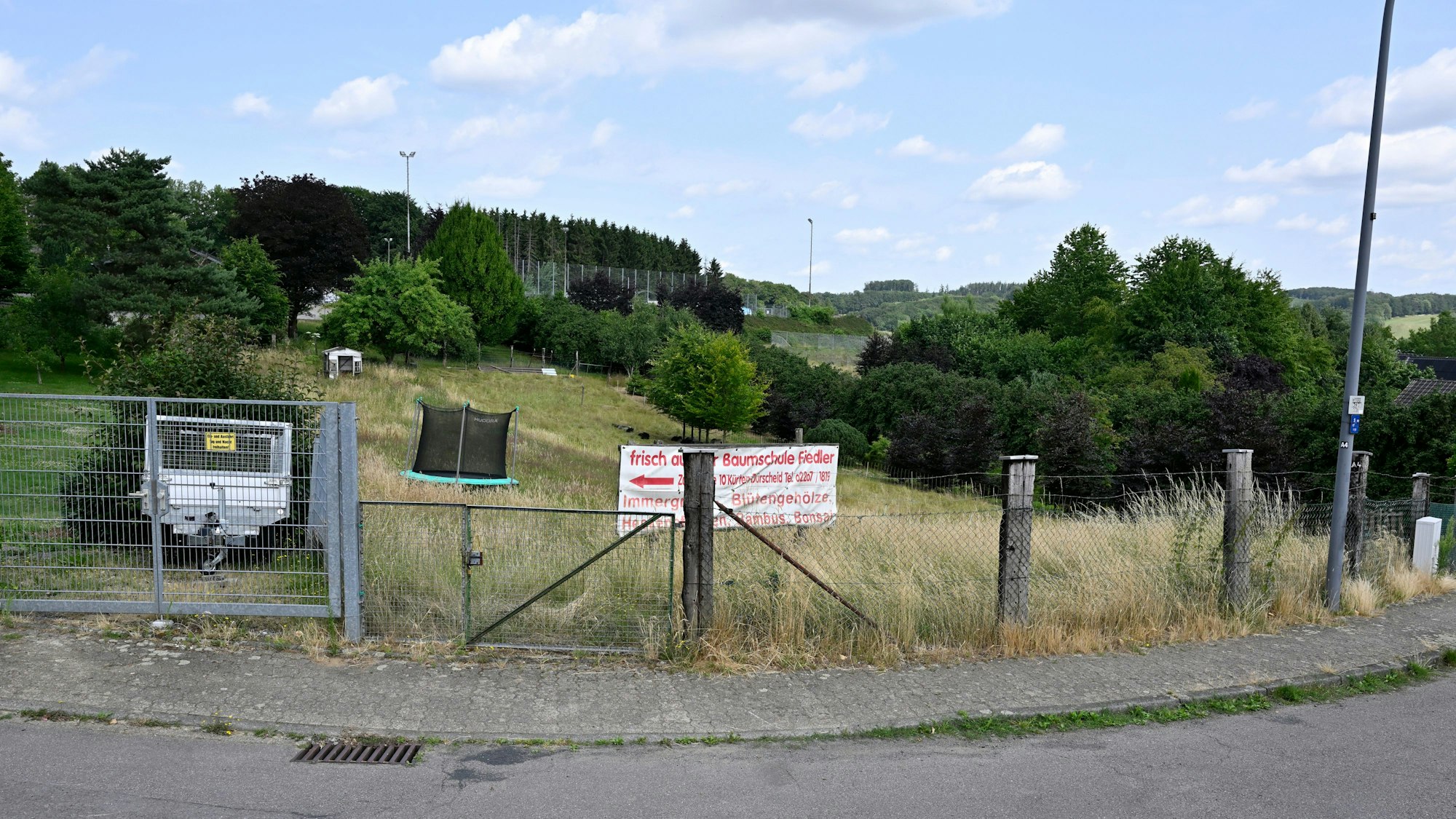 Eine grüne, eingezäunte Fläche mit Werbeschild. Im Hintergrund ein Trampolin.