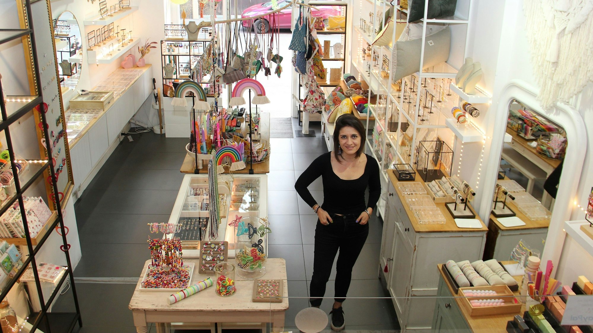 Eine junge Frau mit kinnlangem schwarzem Haar, in schwarzem Shirt und schwarzer Jeans, steht in einem Geschäft mit Tischen und Regalen, in denen Schmuck und Deko-Artikel liegen.