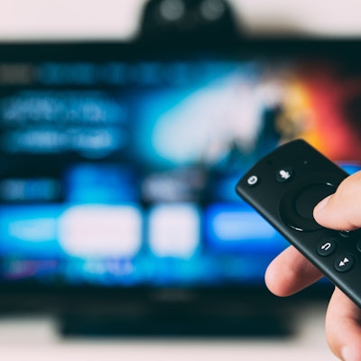 Eine Hand mit einer Fernbedienung vor einem Bildschirm.