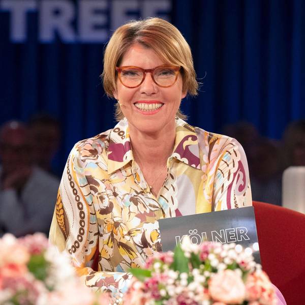 Bettina Böttinger sitzt im Studio des „Kölner Treff“, sie trägt eine buntgemusterte Bluse und lacht in die Kamera.