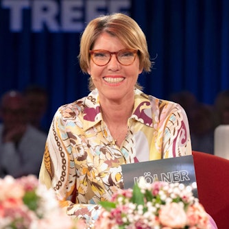 Bettina Böttinger sitzt im Studio des „Kölner Treff“, sie trägt eine buntgemusterte Bluse und lacht in die Kamera.