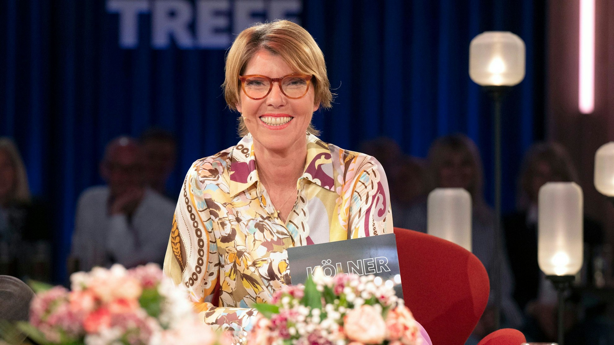 Bettina Böttinger sitzt im Studio des „Kölner Treff“, sie trägt eine buntgemusterte Bluse und lacht in die Kamera.