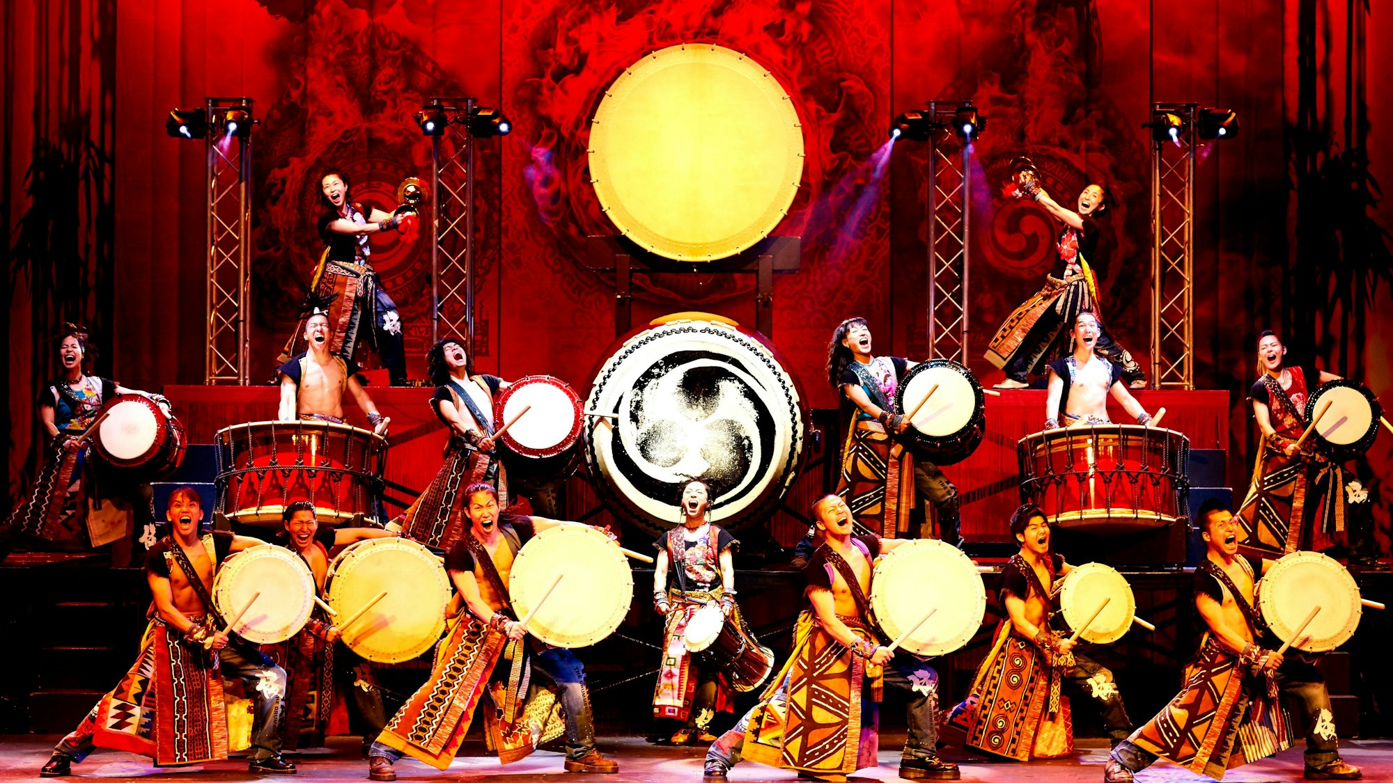 Die Trommler tragen traditionelle Kostüme und tragen ihre Trommeln, die sie an ihre Körper geschnallt haben. Im Hintergrund weitere Trommeln, deren Schlagfläche zur Kamera zeigt. Alles ist in rotes Licht getaucht.