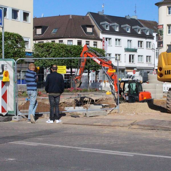 Zwei Männer schauen durch einen Bauzaum auf eine Baustelle. Mehrere Bagger sind zu sehen.