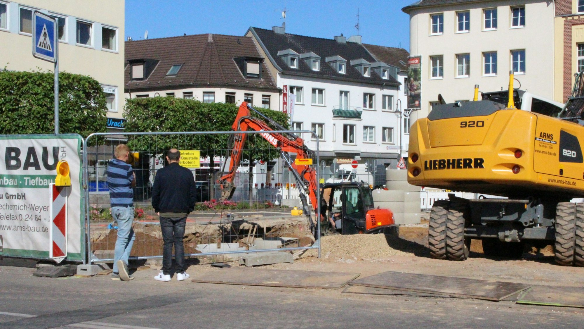 Zwei Männer schauen durch einen Bauzaum auf eine Baustelle. Mehrere Bagger sind zu sehen.