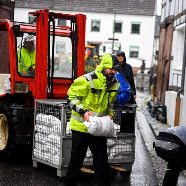 Ein Mann in einer gelben Warnjacke trägt im Regen einen Sandsack von einem Stapel zu einem Tor.