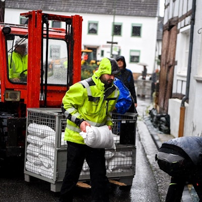 Ein Mann in einer gelben Warnjacke trägt im Regen einen Sandsack von einem Stapel zu einem Tor.