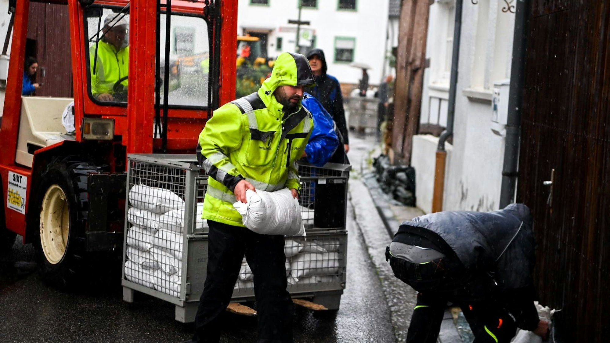 Ein Mann in einer gelben Warnjacke trägt im Regen einen Sandsack von einem Stapel zu einem Tor.