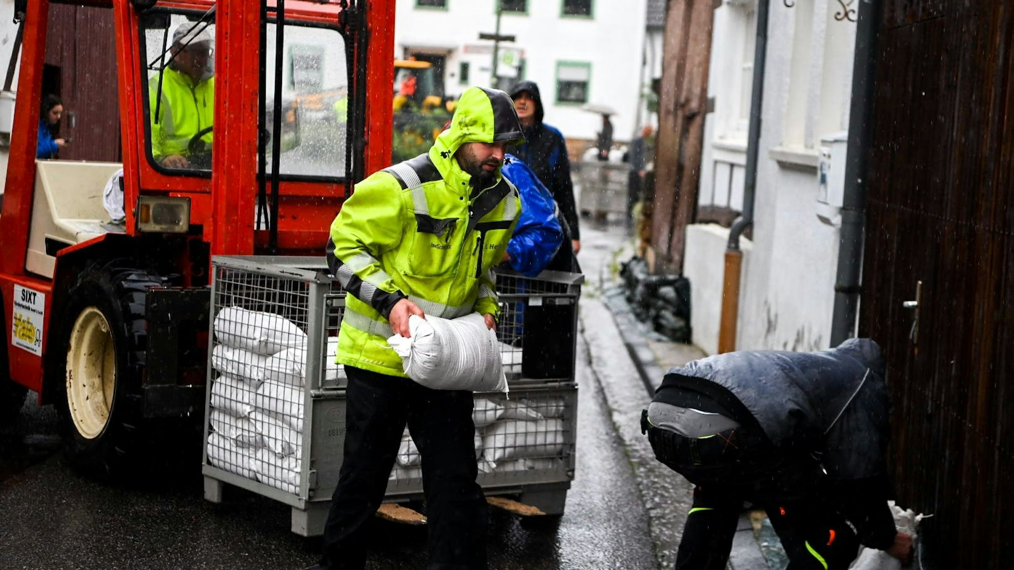 Ein Mann in einer gelben Warnjacke trägt im Regen einen Sandsack von einem Stapel zu einem Tor.