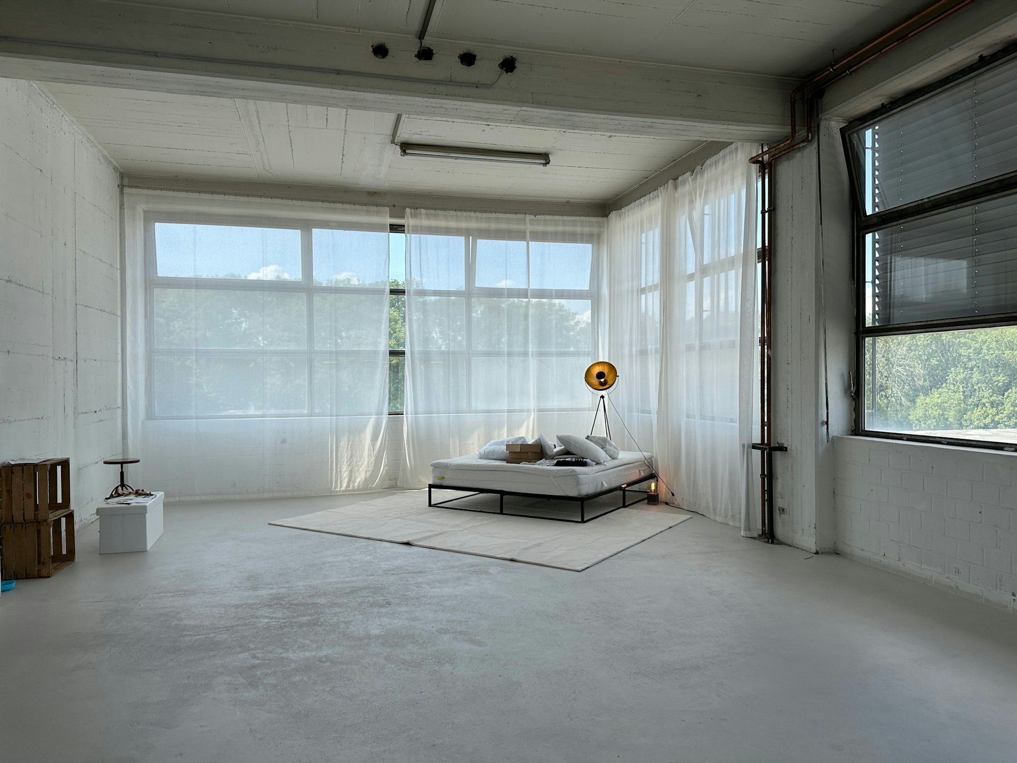 In einem großen Raum mit weißem Betonboden und großen Fenstern steht ein Bett in der Ecke.