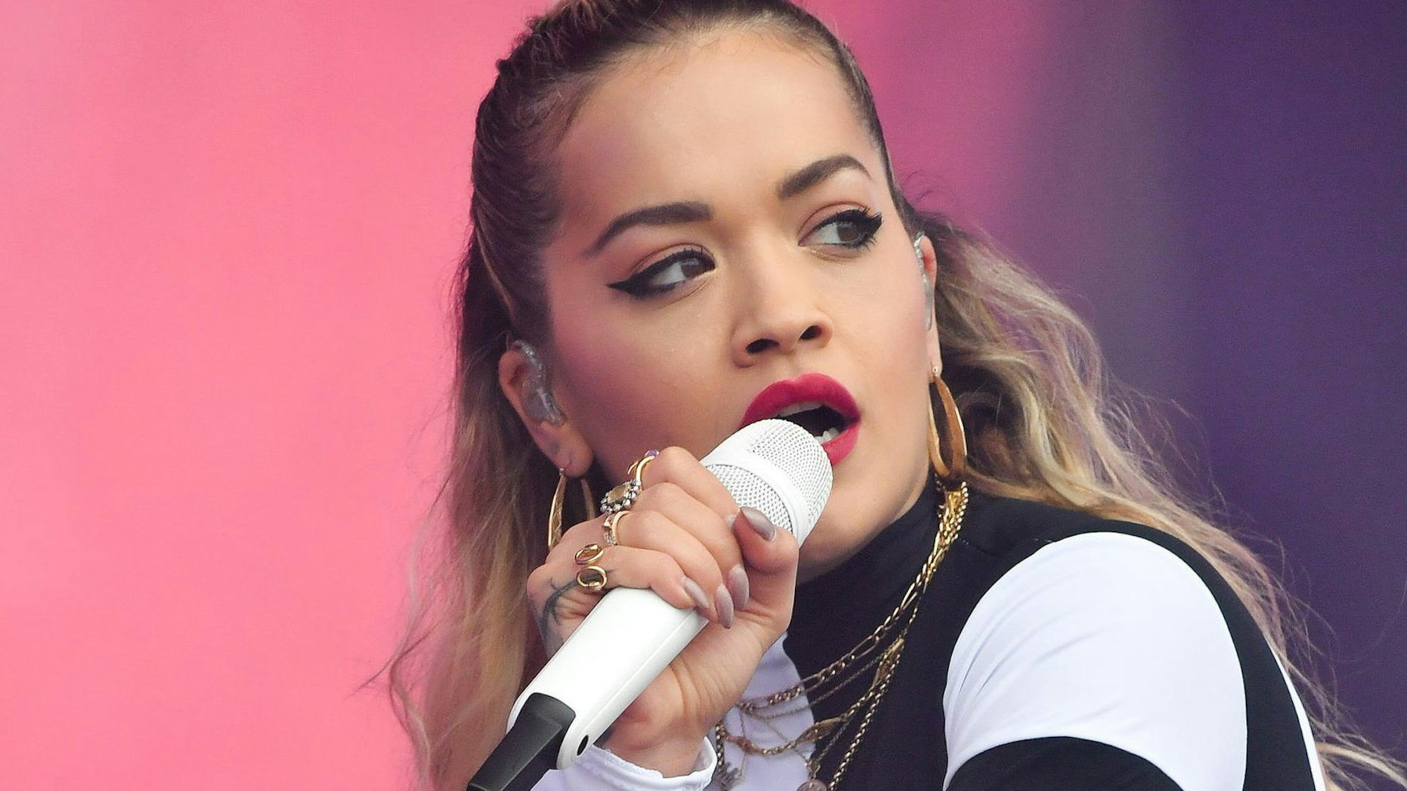 Rita Ora singt in ein weißes Mikrofon, der Hintergrund ist pink und violett.