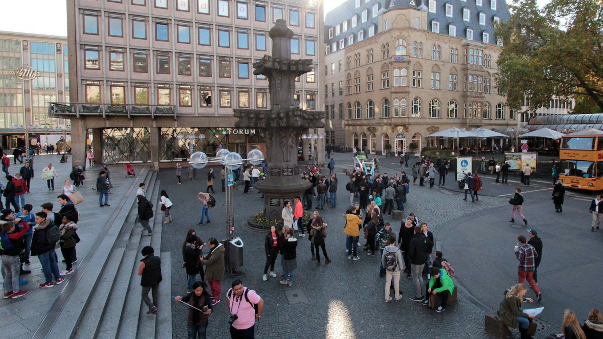 Großer Platz mit darauf stehenden Menschen und einem Brunnen in der Mitte.