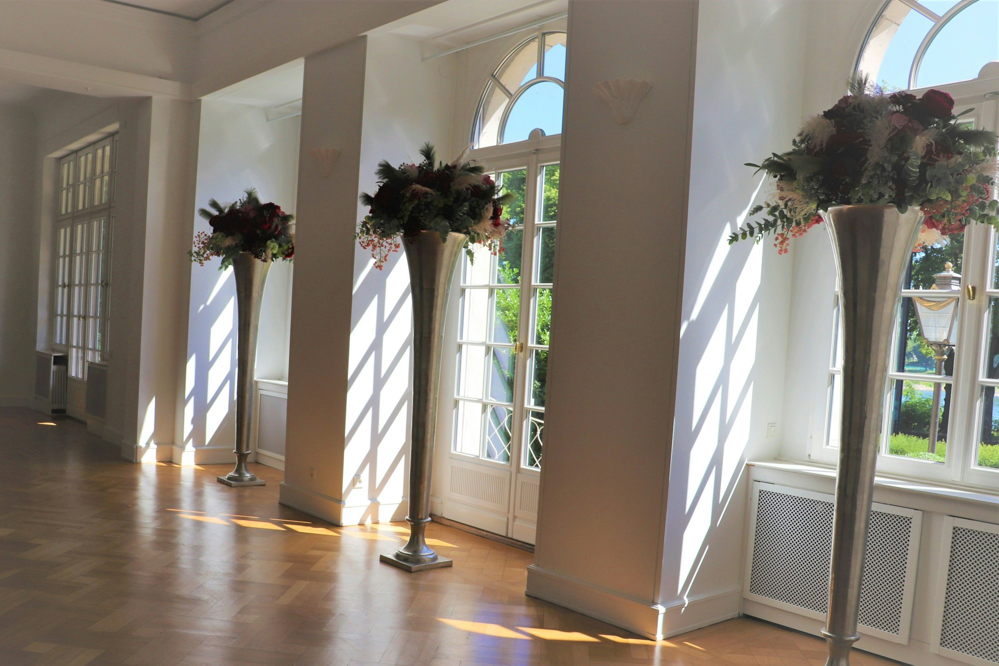 Ein Raum mit Parkett, Terrassentüren und hohen Blumenvasen ist zu sehen.