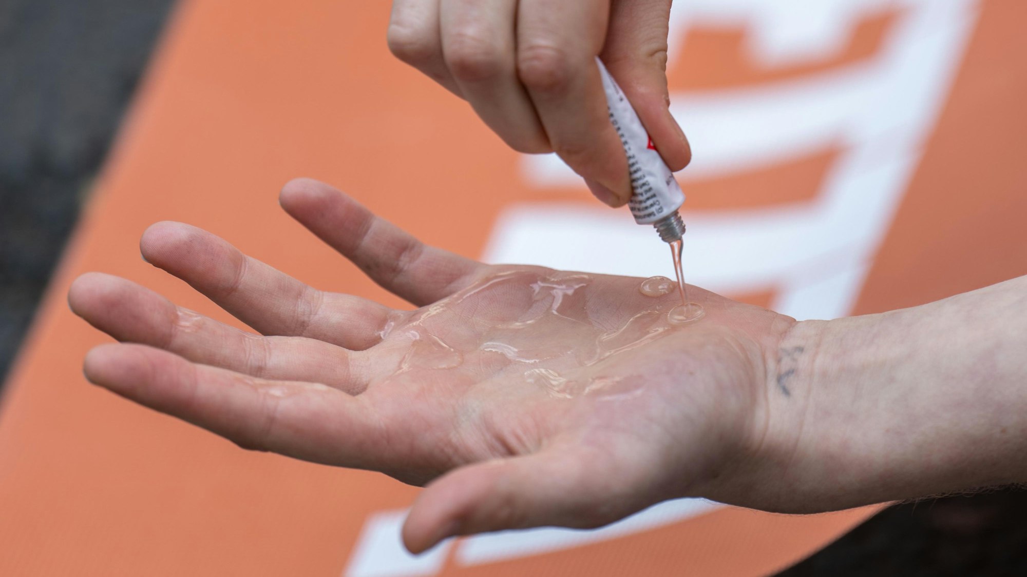 Eine Klimaaktivistin der „Letzten Generation“ schmiert sich Kleber auf die Hand. Im Hintergrund ist ein orangefarbenes Protest-Plakat zu sehen. (Symbolbild)