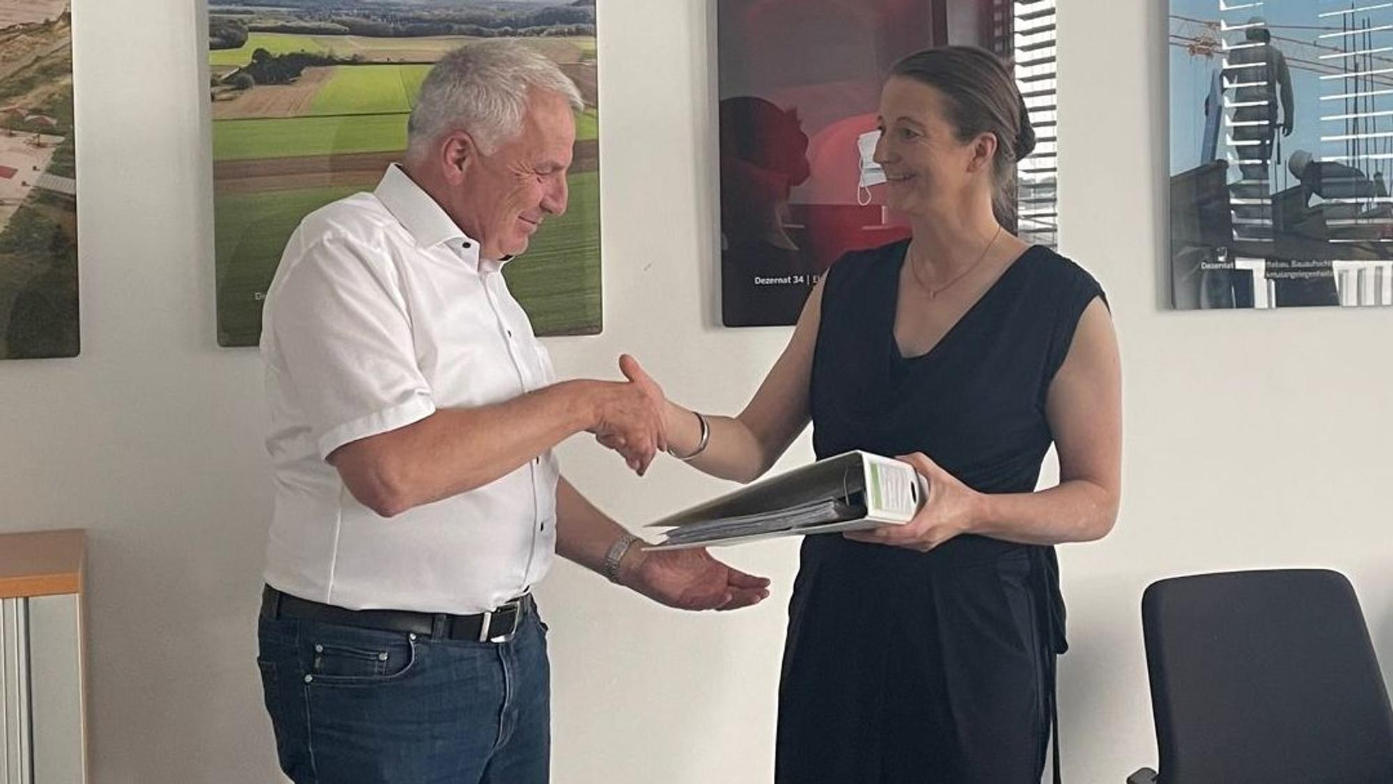 Auf dem Foto sind die Grünen-Politikerin Annika Effertz und Rainer Deppe vom Regionalrat Köln zu sehen. Sie überreicht ihm einen Ordner mit Unterschriften gegen ein geplantes Gewerbegebiet in Kerpen.