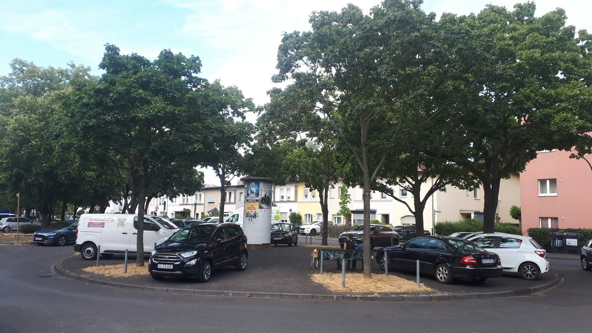 Auf einem Rondell parken Autos, es gibt auch Bäume und eine Litfaßsäule.
