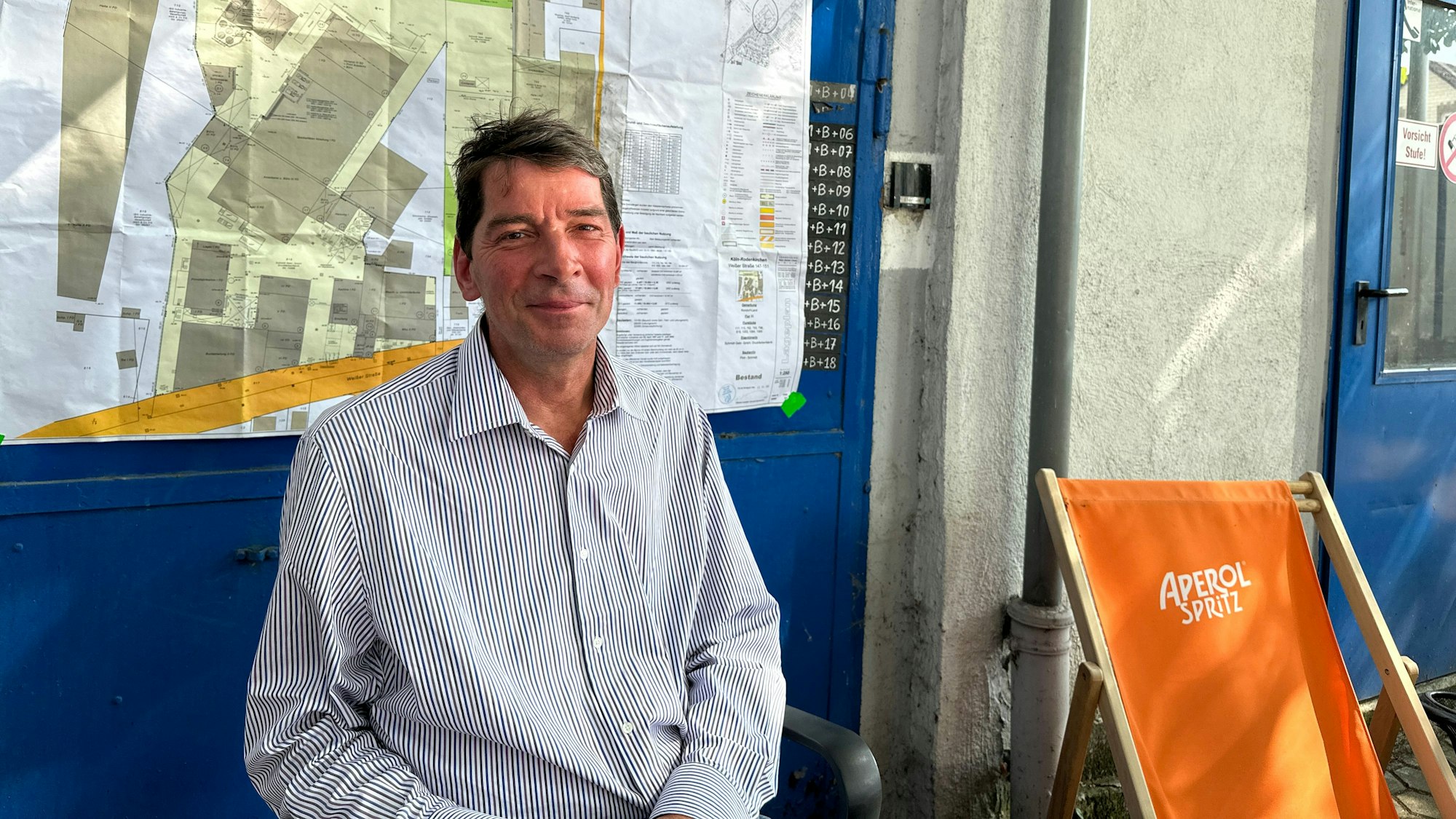 Ein Mann sitzt draußen. Hinter ihm ist ein Lageplan zu sehen.