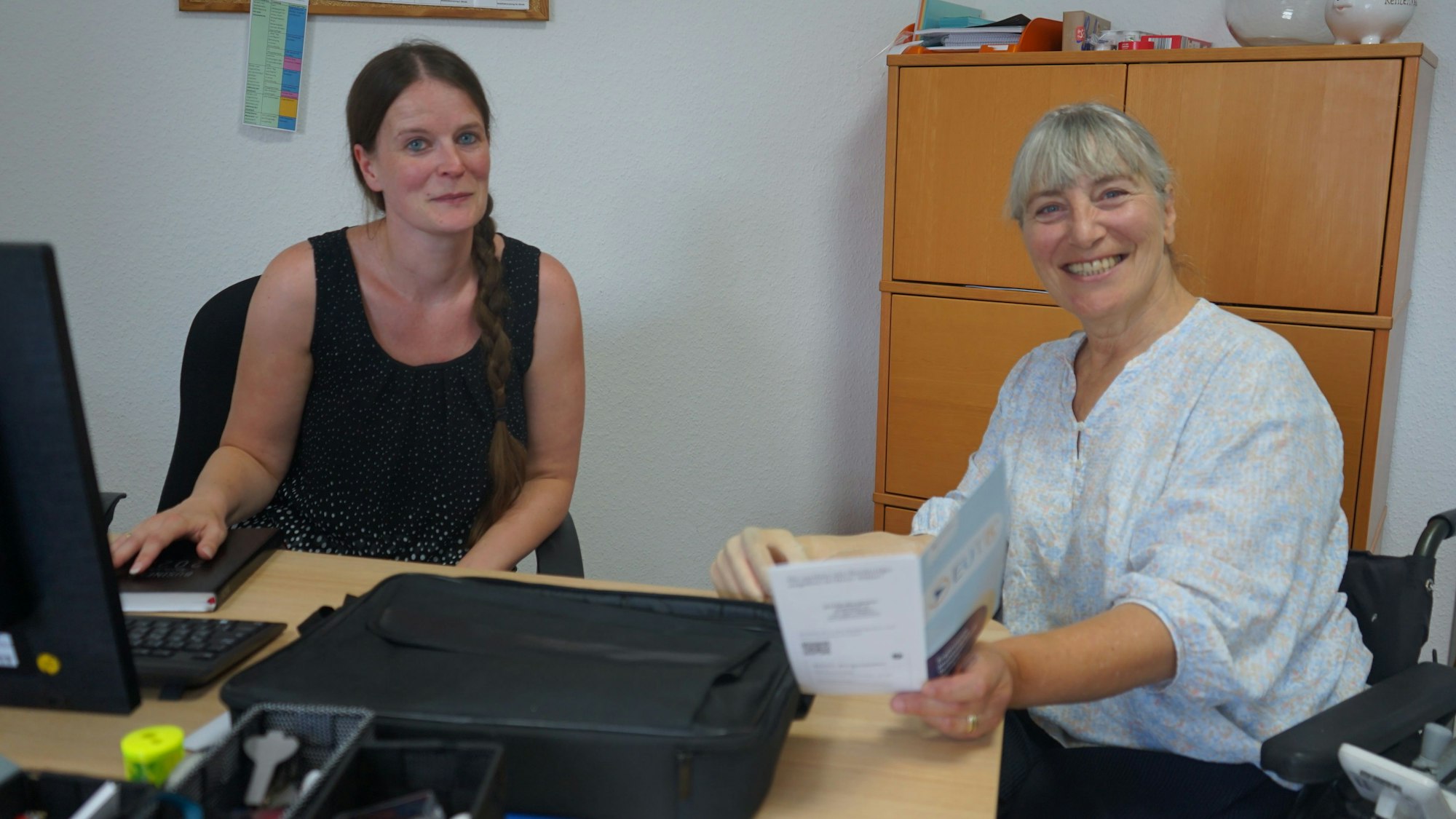 Tanja Langer sitzt am Schreibtisch, vor ihr die Tastatur und ein Notizbuch. rechts daneben sitzt Martina Schankin im Rollstuhl und hält eine Broschüre in ihrer Hand.