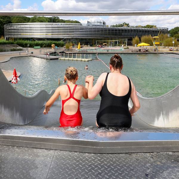 Das Bild zeigt eine Mutter und ihre Tochter von hinten, die einen Wasserrutsche hinunterrutschen. Im Hintergrund sieht man den Naturbadeteich im Lentpark Freibad in Köln.