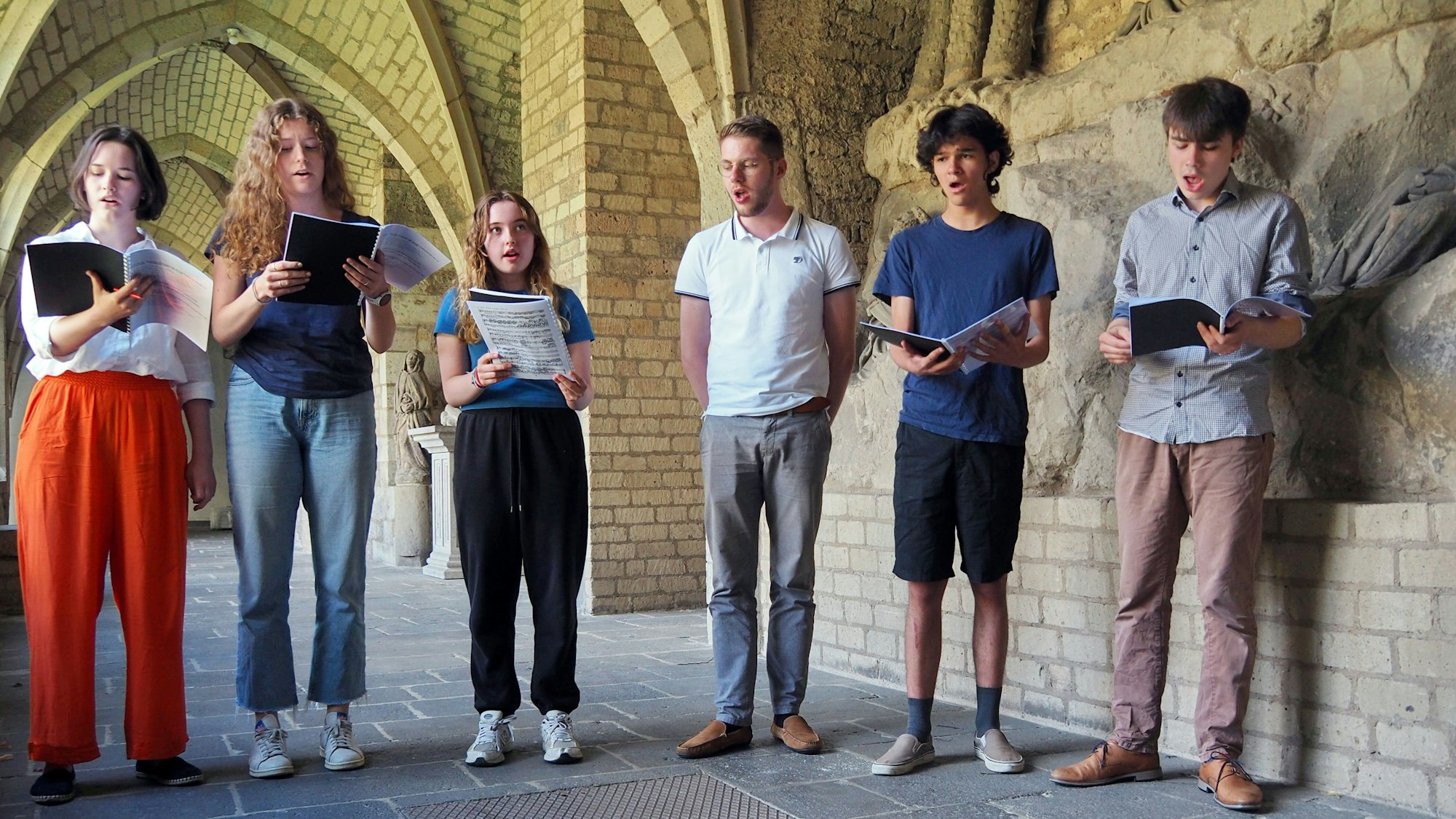 Drei Mädchen und drei Jungs stehen nebeneinander in einem Kreuzgang und singen vom Blatt.