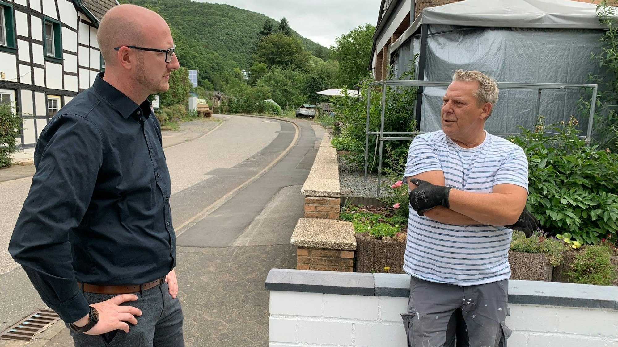 Jürgen Dopperath lehnt an einer hüfthohen Mauer. Er spricht mit Bürgermeister Ingo Pfennings und berichtet vom Wiederaufbau seines Hauses.