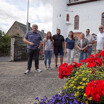 Eine Gruppe von Menschen steht auf einem Platz. Im Hintergrund ist ein Teil einer Kirche zu erkennen, im Vordergrund stehen gelbe, rote und lila Blumen.