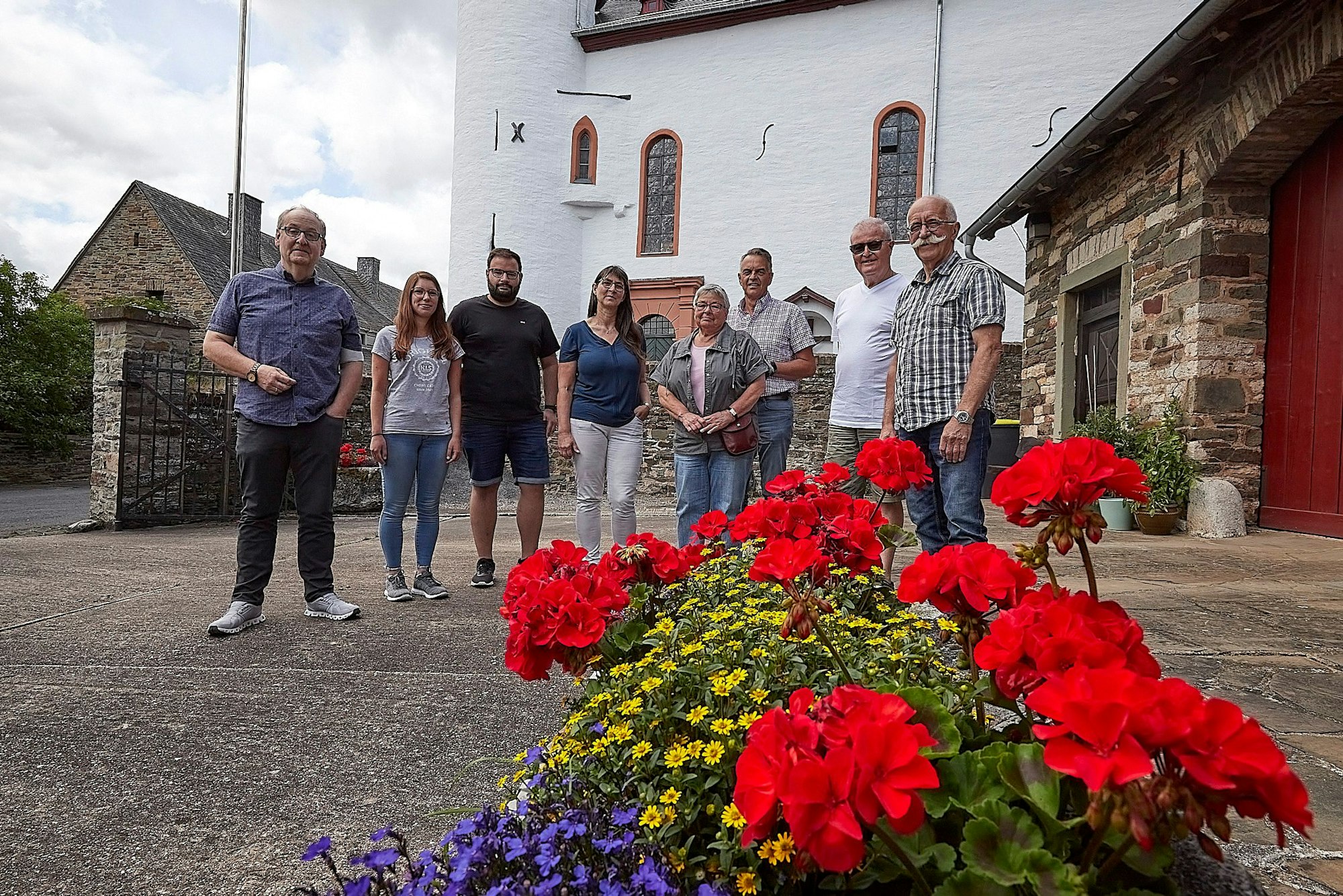 Eine Gruppe von Menschen steht auf einem Platz. Im Hintergrund ist ein Teil einer Kirche zu erkennen, im Vordergrund stehen gelbe, rote und lila Blumen.