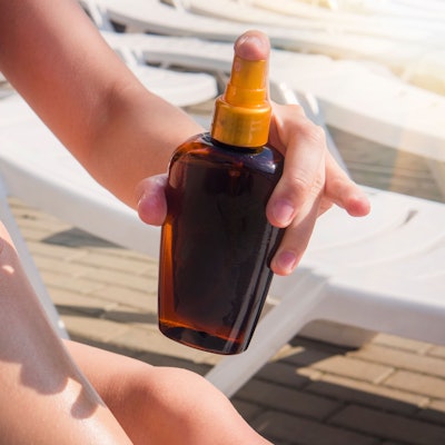 Eine Frau sitzt auf einer Sonnenliege und sprüht Bräunungsöl auf ihr Bein.