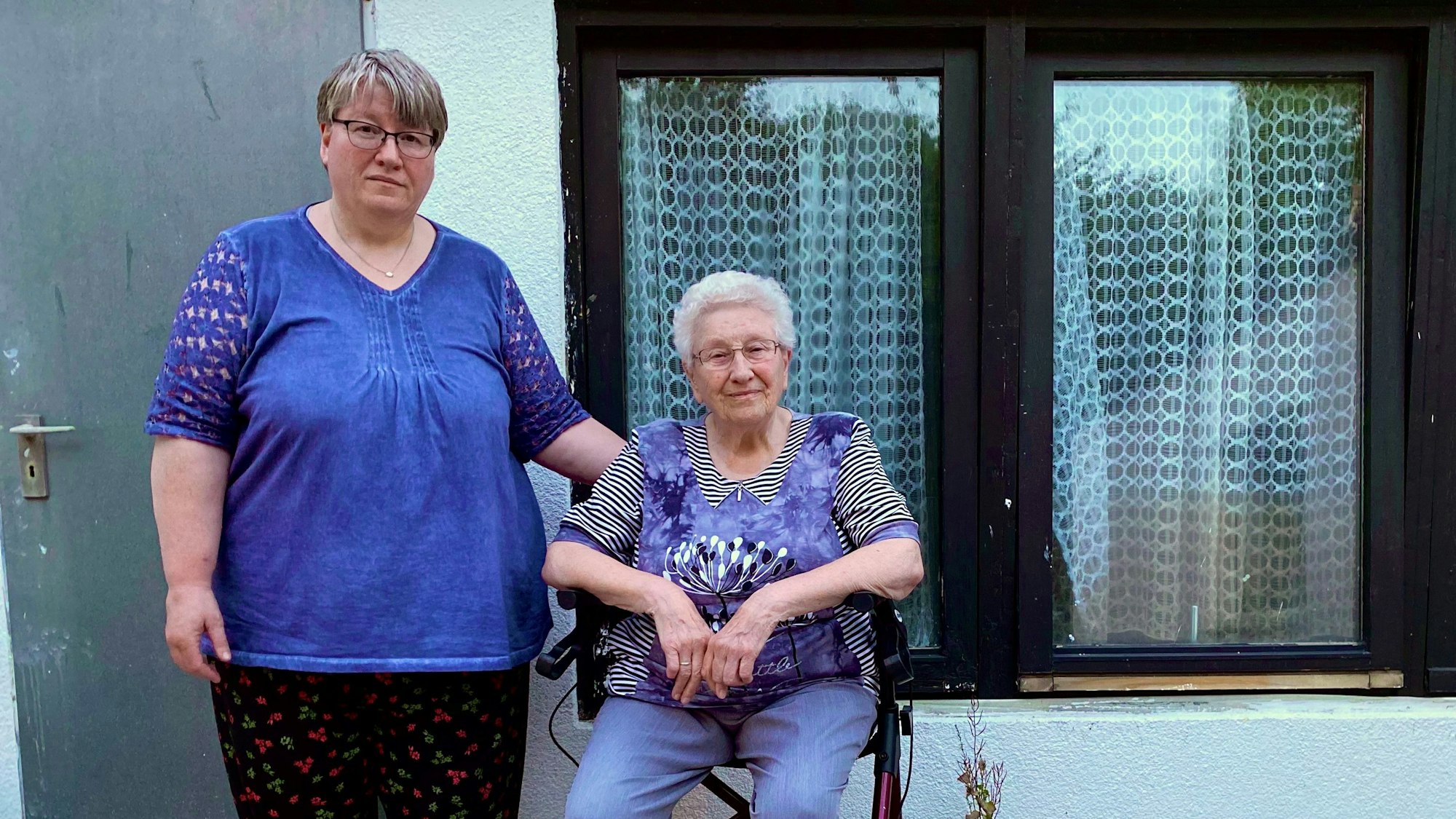 Claudia Carls steht und Marianne Carls sitzt vor ihrem Partyraum in Euskirchen-Roitzheim. An den Gardinen ist durch braune Einfärbung zu erkennen, bis wohin das Wasser stand.