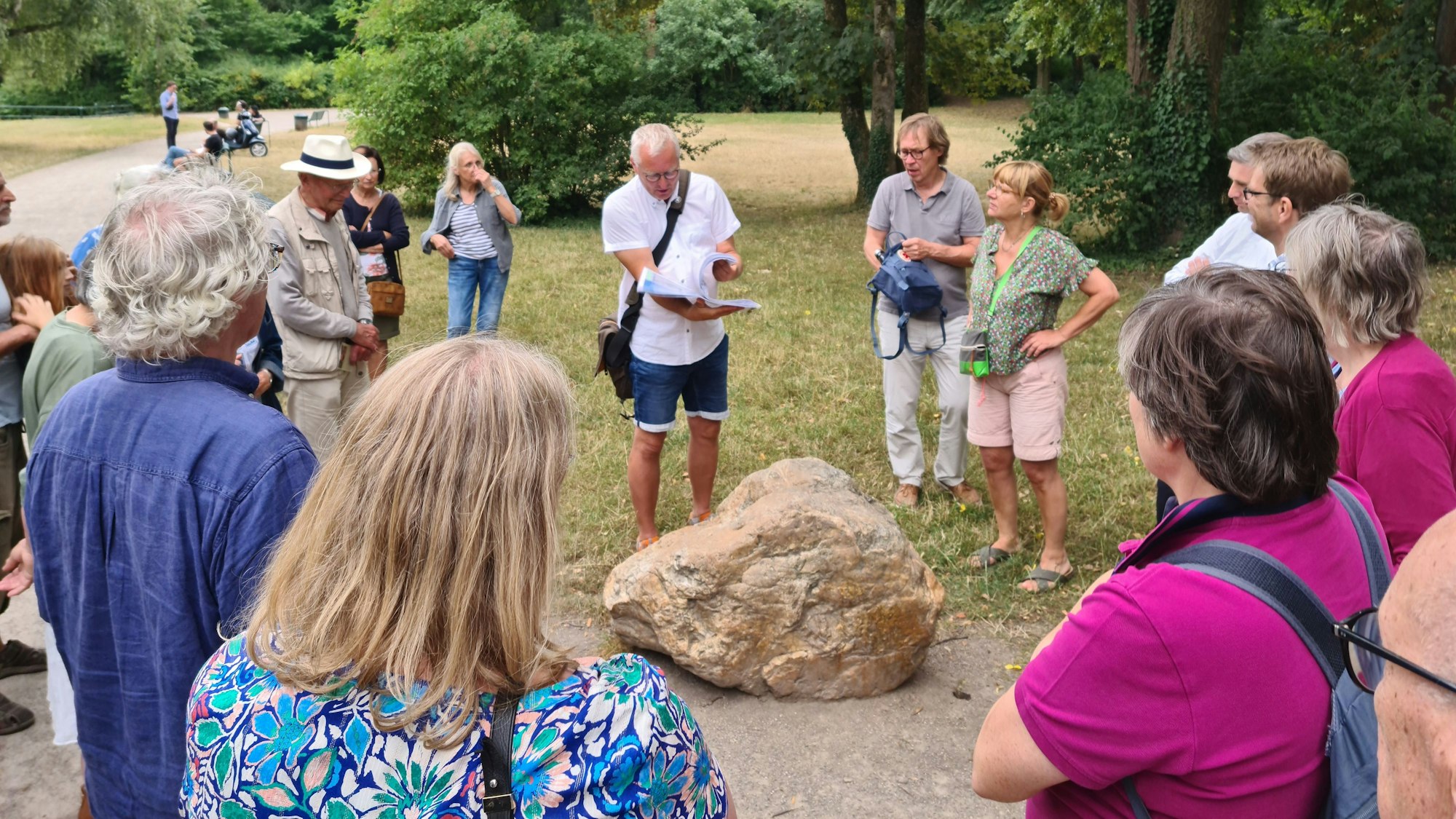 Eine Gruppe Menschen steht im Halbkreis um einen Mann im weißen Hemd, der eine Welle formt aus einer Mappe mit geologischen Karten.