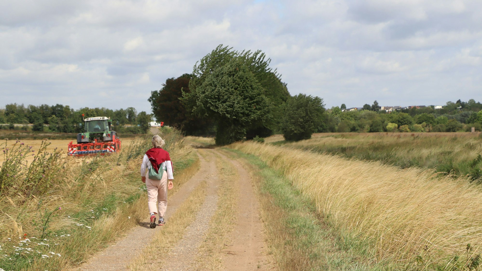 Eine Frau wandert zwischen Felder, auf dem Acker fährt ein Traktor.