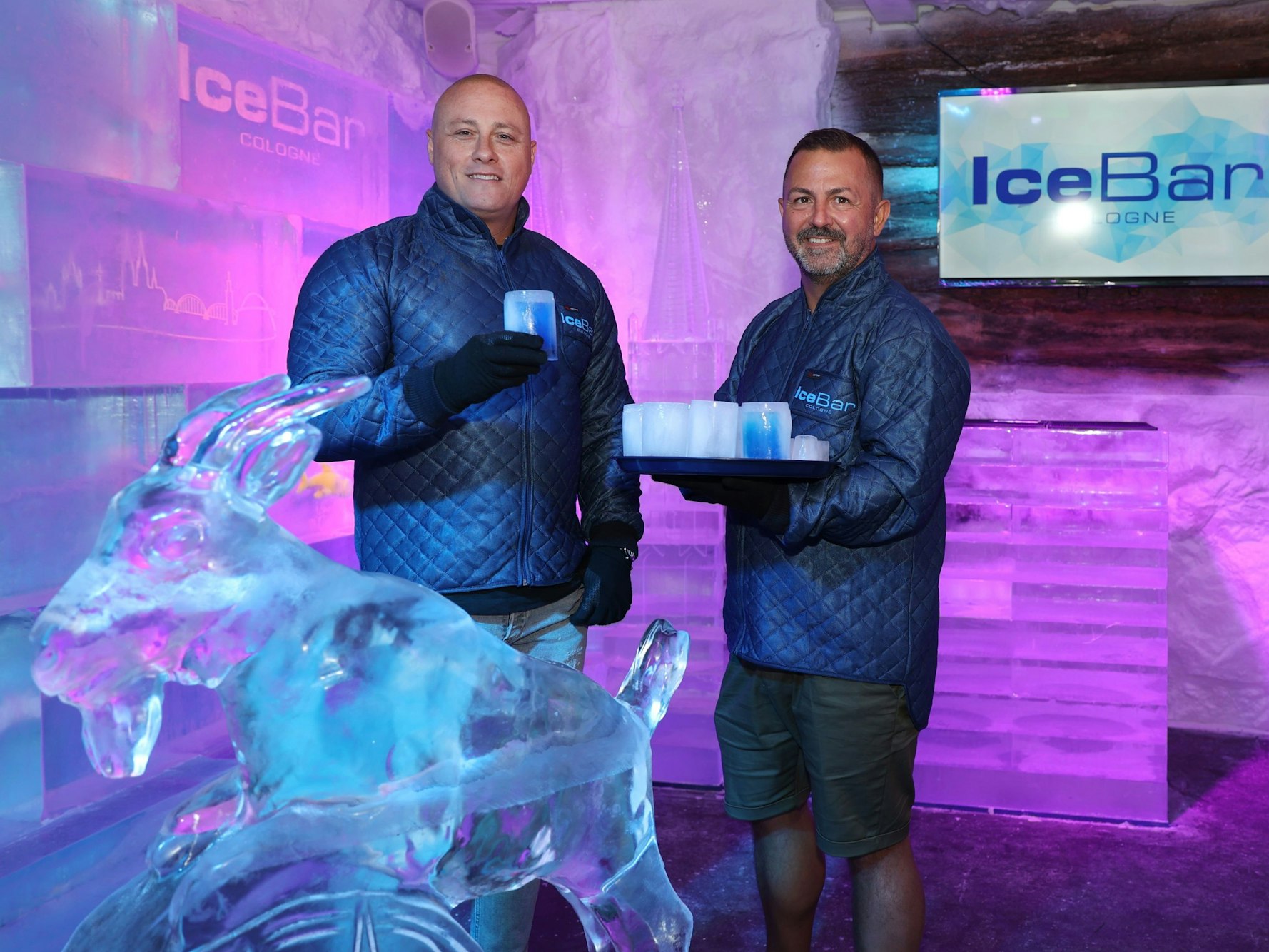 Markus Weiss (links) und Stefano Straberg in der Kältekammer der „Ice Bar Cologne", einem Raum aus Eis. Vor ihnen ein Geißbock aus Eis, hinter ihnen ein Logo. Sie halten Gläser aus Eis.