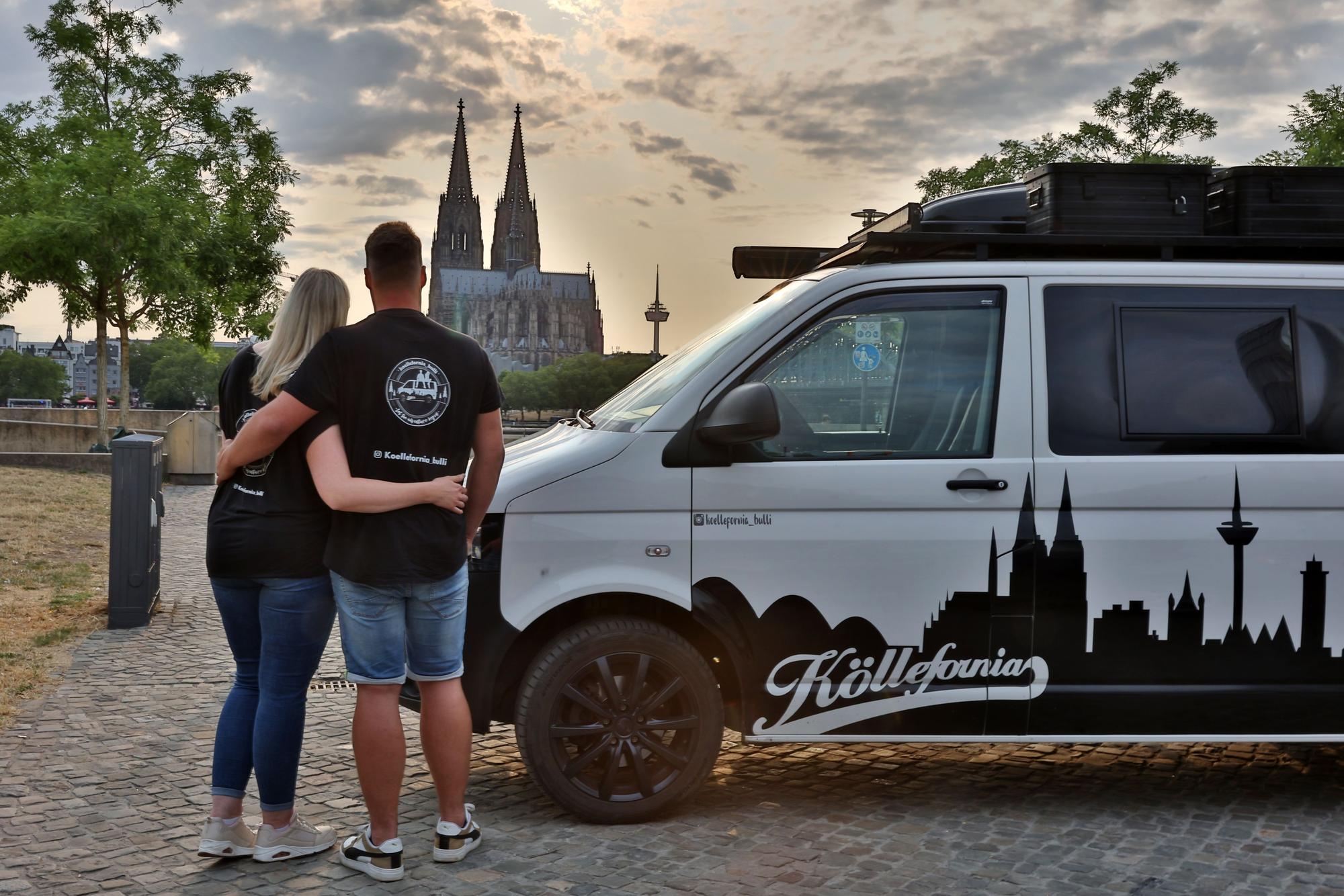 Zwei Menschen von hinten stehen neben dem VW Transporter. Im Hintergrund ist der Kölner Dom bei Sonnenuntergang zu sehen.
