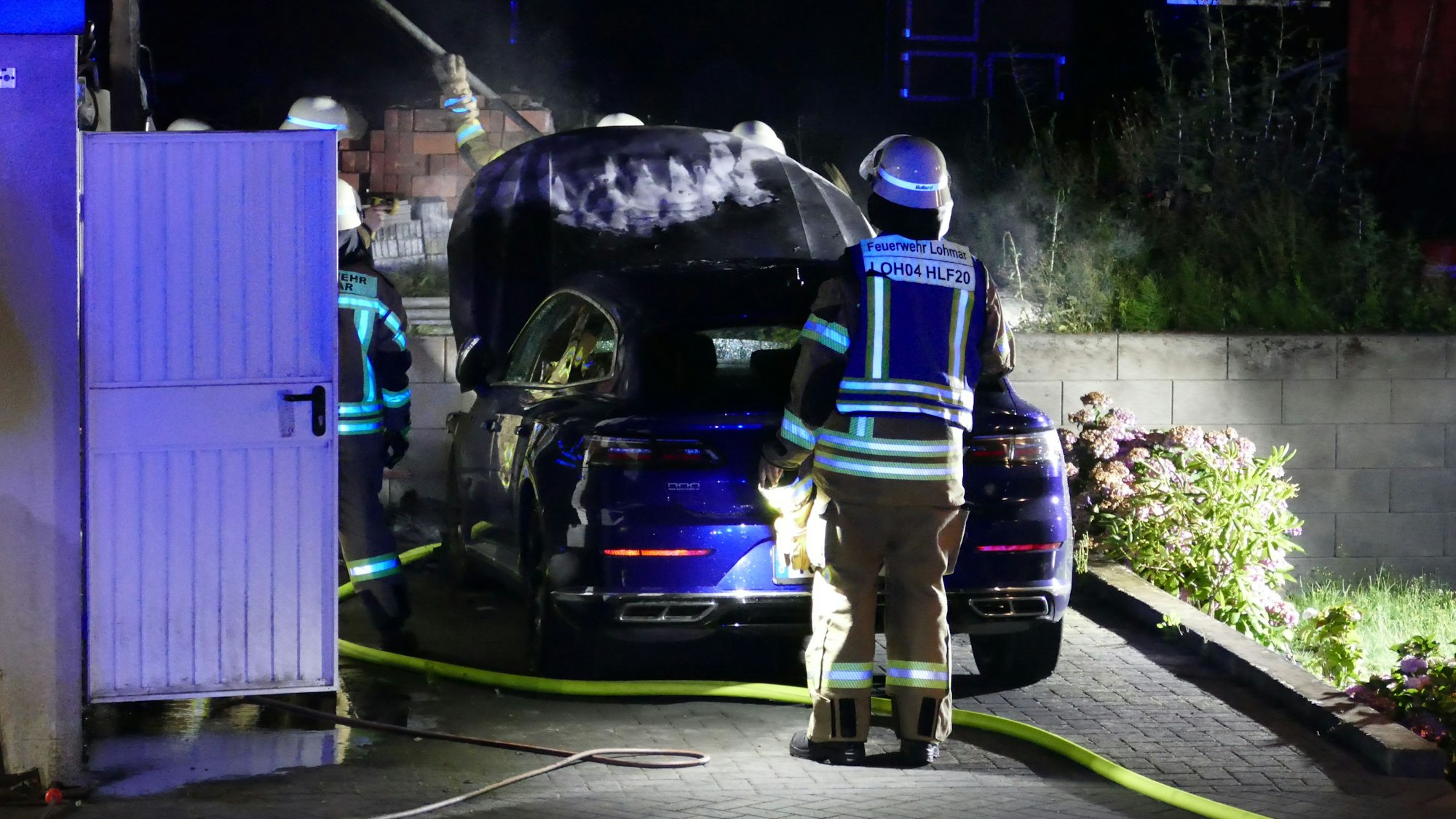 Man sieht eine Person in Feuerwehrkleidung vor einem Auto stehen, die Motorhaube ist offen, ein Löschschlauch liegt auf dem Boden.