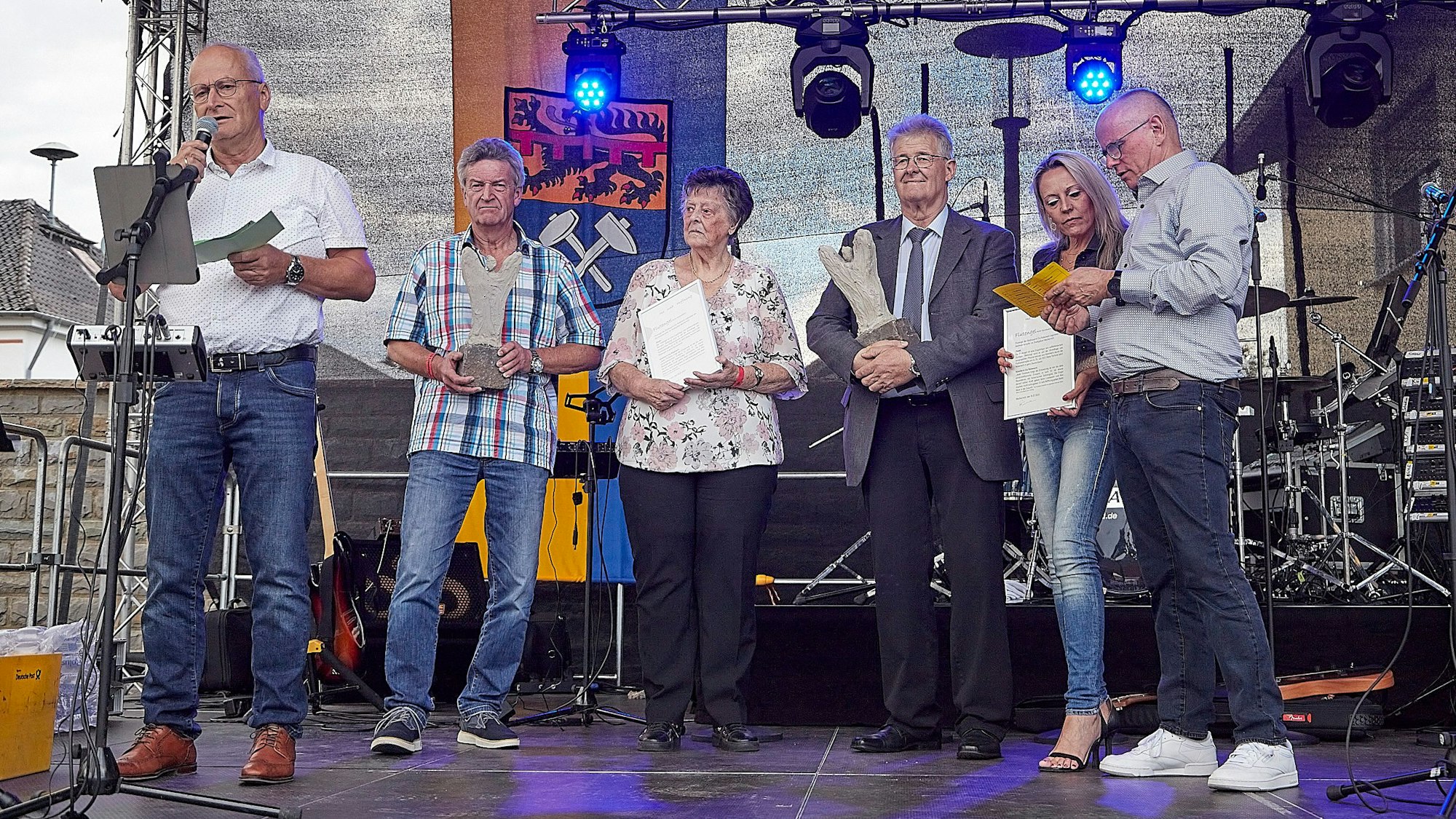 Mehrere Menschen stehen auf einer Bühne, einer spricht über ein Mikrofon zum Publikum.